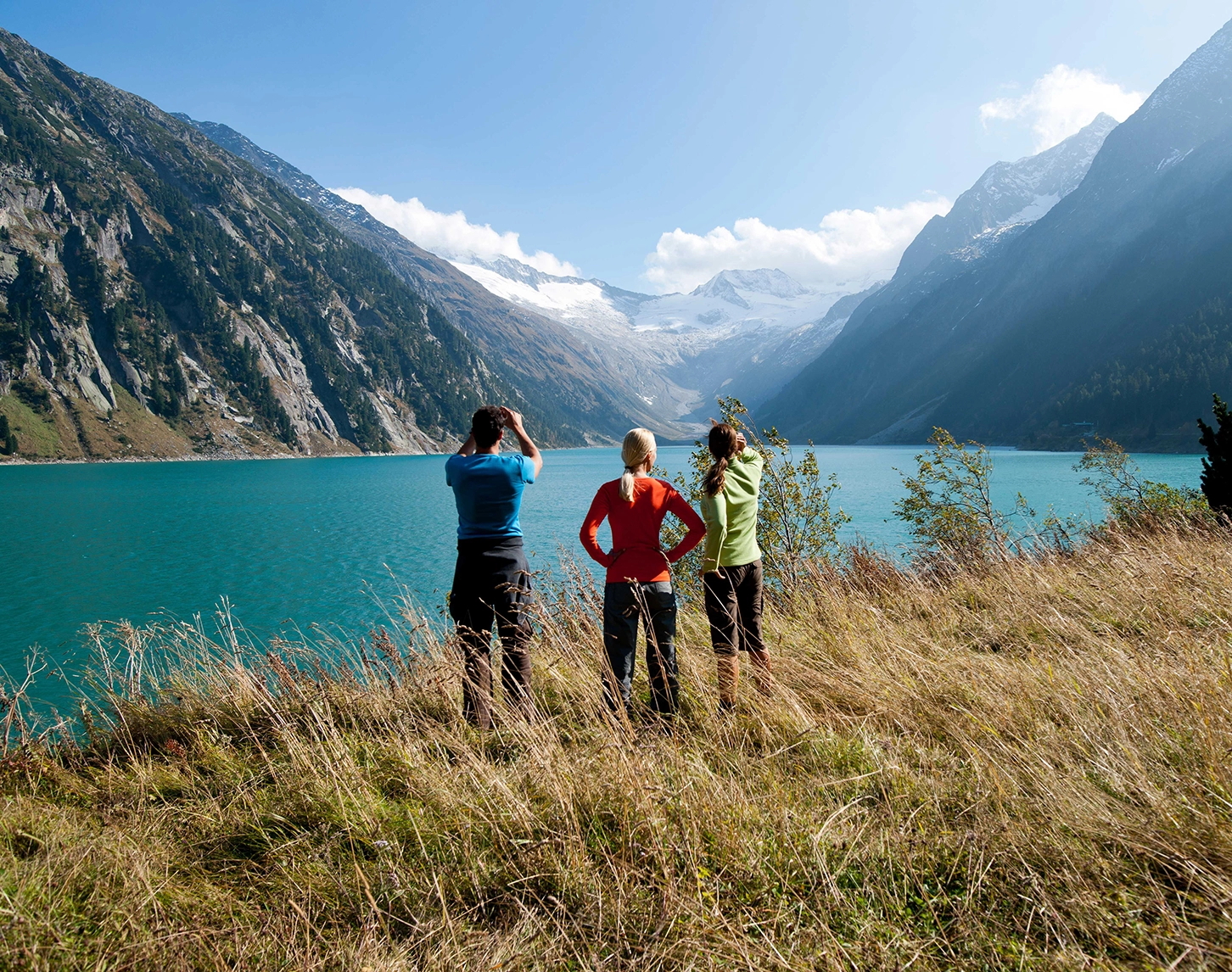 Drei Wanderer stehen auf einer etwas bräunlichen Bergwiese am Stausee in Schlegeis. Wir sehen nur ihren Rückenansicht. Im Hintergrund ist der See zu sehen und die Alpen. Der Himmel ist blau.