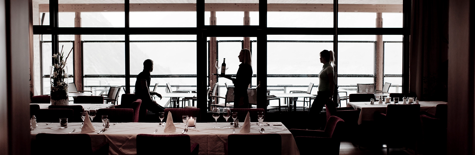 Wir befinden uns im Inneren des Berghotel Maltas. Die Aufnahme zeigt das Restaurant. Einige Mitarbeit sind im Gastraum damit beschäftig diesen für die Gäste herzurichten. Das Bild wurde gegen das Sonnenlicht aufgenommen, dadurch entsteht eine Art Scherenschnitt mit starken schwarzen Kontrasten.