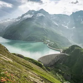 Wir blicken von oben auf den Stausee Mooserboden in Kaprun. Die Stimmung ist frühlingshaft. Ein leichter Nebel schwebt über dem See. Die Sonnenstrahlen leuchten dazwischen.