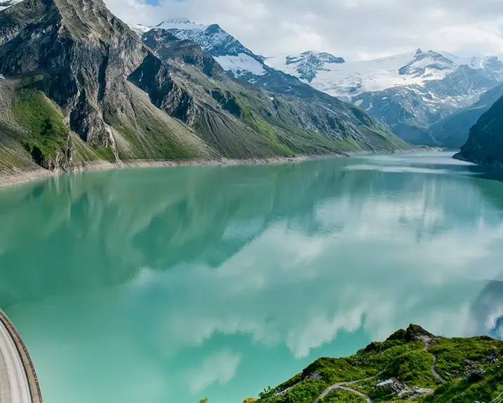 Wir blicken auf den Stausee Mooserboden in Kaprun. Berge und Wolken spiegeln sich im türkisfarbenen Wasser wieder.